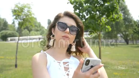 时尚迷人的女孩看着手机在一个城市公园里梳头视频的预览图