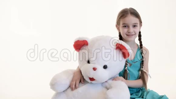 一个可爱的白色女孩的肖像她扎着辫子抱着一只大泰迪熊坐在地板上背景是白色的视频的预览图