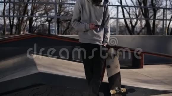 阳光明媚的一天年轻卷曲的头男滑板运动员在滑板公园里玩手机靠在栏杆上视频的预览图