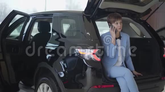 自信的女人短发穿着经典的蓝色正式西装坐在敞篷车后备箱里用手机说话女孩视频的预览图