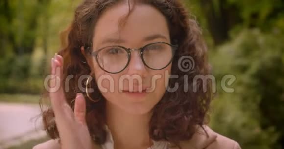 年轻漂亮的长发卷曲的高加索女性戴眼镜的特写照片微笑着愉快地看着镜头视频的预览图