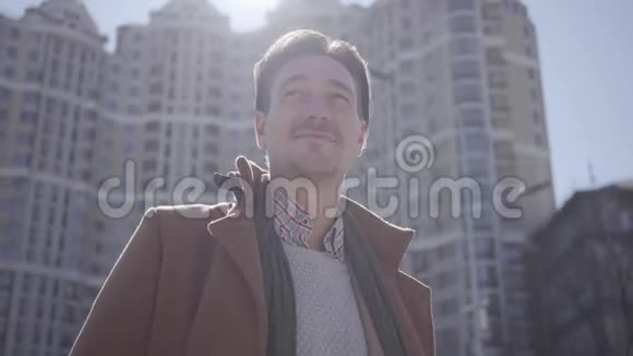 微笑的英俊自信的男人穿着棕色外套站在城市街道上环顾四周的肖像高层建筑视频的预览图