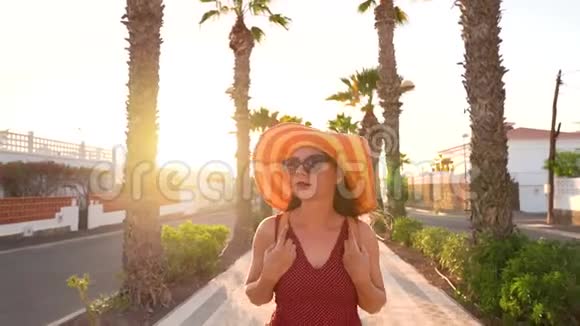 日落时分戴着一顶黄色大帽子的快乐女人沿着棕榈巷奔跑慢动作视频的预览图