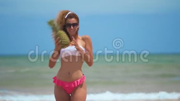 有趣的微笑女孩在夏季泳装和太阳镜性感的女人带着新鲜的菠萝在附近积极跳舞视频的预览图