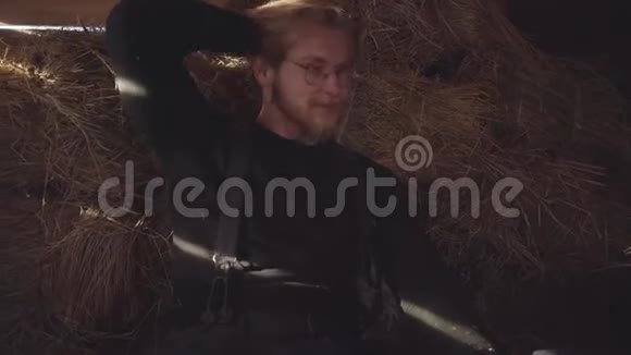 一个英俊的留胡子的男人戴着眼镜坐在马厩里的干草上的肖像那家伙从毛衣上抖干草视频的预览图