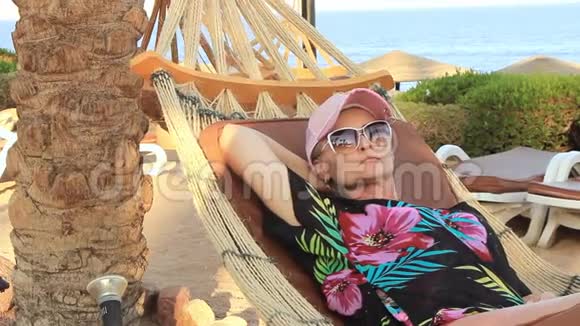 美丽的年轻女子戴着帽子在阳光明媚的夏日棕榈树的树荫下躺在海滨的吊床上视频的预览图