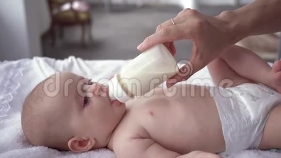 妈妈照顾穿着尿布的女婴躺在换尿布的桌子上喝着妈妈拿在里面的瓶子里的牛奶视频的预览图