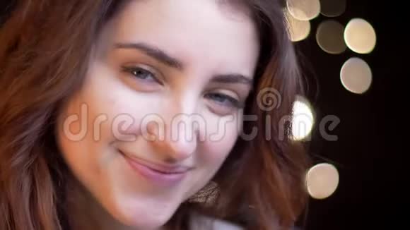 在模糊的灯光背景下年轻的白种人女孩在镜头中微笑的特写肖像视频的预览图