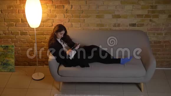 年轻的白种人女孩波浪状的头发躺在沙发上在舒适的家庭氛围中专注地看着平板电脑视频的预览图