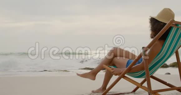 在海滩上的阳光休息室里放松的非裔美国妇女的侧面景色视频的预览图