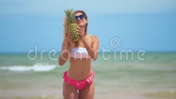 有趣的微笑女孩在夏季泳装和太阳镜性感的女人带着新鲜的菠萝在附近积极跳舞视频的预览图