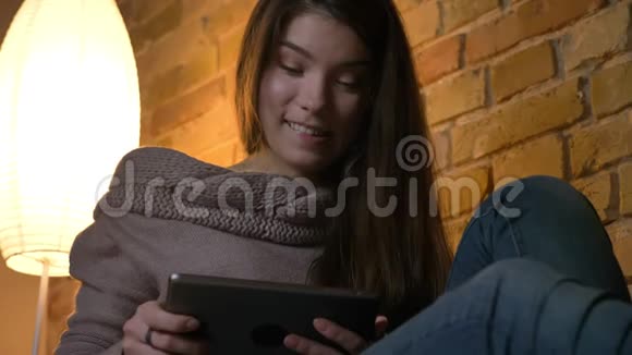 从底部的年轻白种人女孩坐在沙发上沉思地看着平板电脑舒适的家庭背景视频的预览图