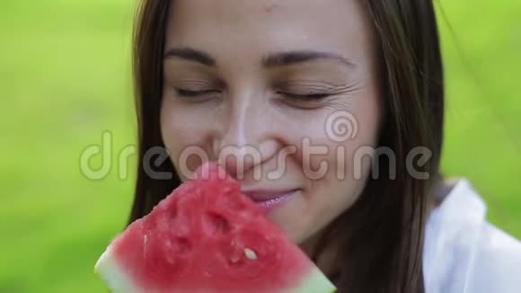 特写女孩的肖像闻着香气吃着一个红色的健康多汁的西瓜在绿色的西瓜上傻笑视频的预览图