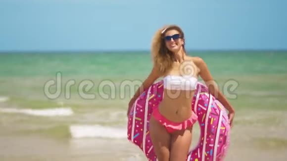 女人玩得开心笑着和粉红色的甜甜圈跳舞穿比基尼的女孩在海滩上戴太阳镜美丽性感的女人视频的预览图