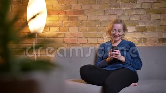 高白种人女性使用手机的特写镜头笑着坐在室内的沙发上视频的预览图