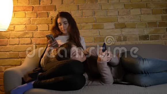 年轻的高加索女孩在沙发上放松在舒适的家庭氛围中观看他们的智能手机的肖像视频的预览图