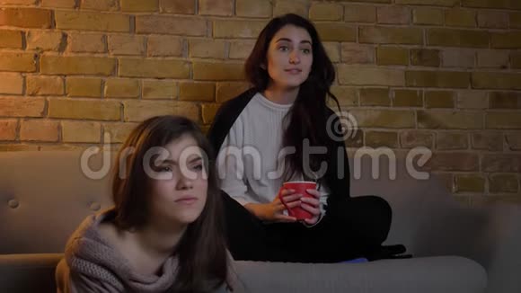 特写两个年轻的白种人女孩坐在客厅里用不同的情绪看电视视频的预览图