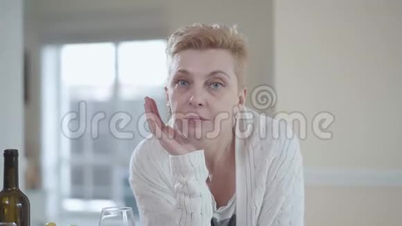 穿着白色毛衣的悲伤老年妇女的肖像靠在桌子上在室内近距离看着镜头背景资料视频的预览图