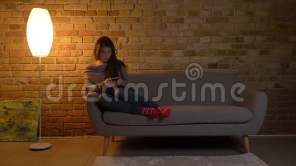 年轻的白种人女孩坐在沙发上在舒适的家庭背景下微笑地看着智能手机的肖像视频的预览图