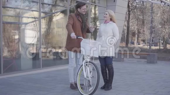 高个男人拿着自行车和穿着暖和夹克的金发美女聊天一对积极的夫妇站在视频的预览图