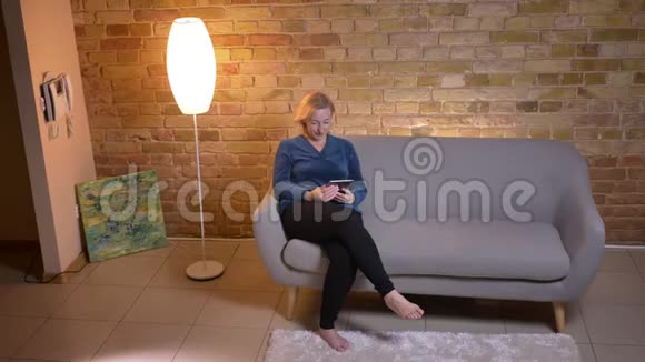 一位高水平的高加索女人在平板电脑上发短信坐在舒适的沙发上开心地笑了视频的预览图