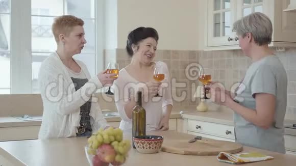 三位积极的成熟女性在家中交流靠近厨房的现代餐桌一位女士说祝酒词他们叮当作响视频的预览图