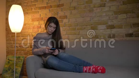 集中的白种人女孩坐在沙发上严肃地看着智能手机在舒适的家里看着相机视频的预览图