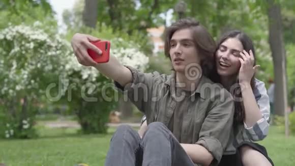 一对可爱的学生坐在公园的草地上自拍他在前台女孩拥抱视频的预览图