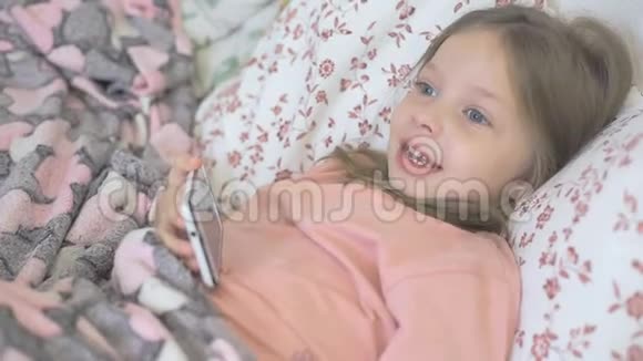 一个生病的小女孩和她的母亲躺在床上看着智能手机屏幕上的卡通片儿童和数字视频的预览图
