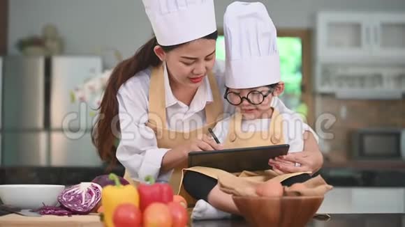 漂亮的亚洲女人和带眼镜的可爱的小男孩准备在家里的厨房里用平板电脑做饭人民的生活方式和视频的预览图