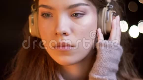 戴着耳机听着模糊灯光下的音乐的年轻美丽的高加索女孩的特写肖像视频的预览图