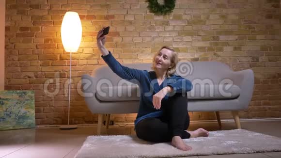 在一间舒适的公寓里一位年长的白种人女性坐在地板上一边打电话一边自拍特写镜头视频的预览图