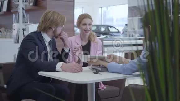 未被识别的汽车经销商穿着时髦的西装在平板电脑上向年轻的成功夫妇展示他们的新车的信息视频的预览图