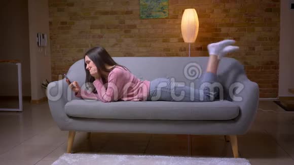一位年轻漂亮的黑发白种人女性在沙发上躺在一个舒适的沙发上用电话和微笑特写镜头视频的预览图