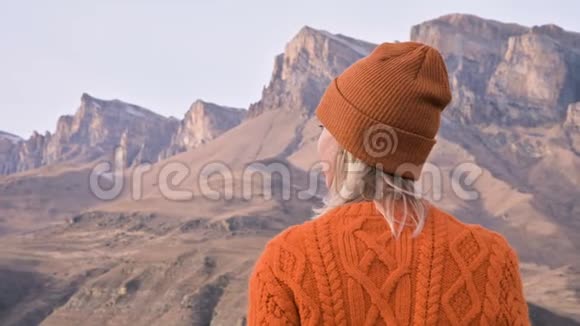 从后面看一个穿着橙色毛衣和帽子的旅行女孩在户外看台上以史诗般的背景为背景视频的预览图