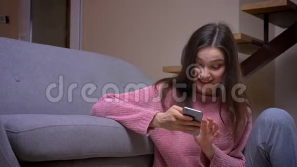 一位年轻漂亮的褐发白种人女性在一个舒适的房间里坐在地板上用电话和微笑拍摄特写镜头视频的预览图