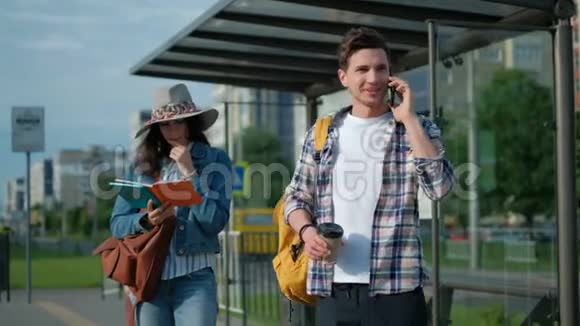 年轻人和女孩站在公共汽车站太阳照耀着他在用手机在等他的电车城市视频的预览图