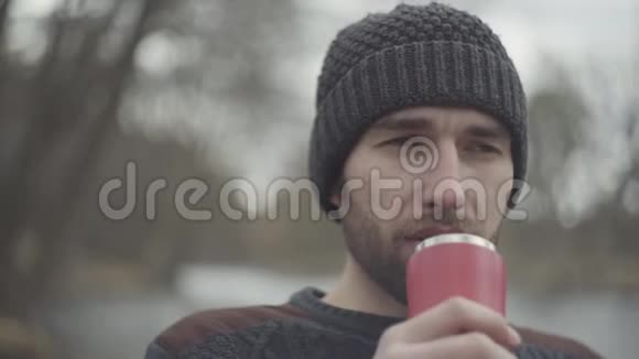 一个留着胡子自信微笑的时髦男人戴着温暖的帽子在户外喝茶或喝咖啡的肖像这就是视频的预览图