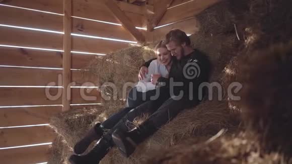 戴眼镜的长胡子的英俊男人和没有暖布的漂亮女人坐在寒冷的冬天的谷仓里的干草上青年视频的预览图