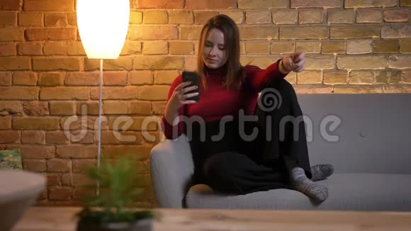 年轻漂亮的白种人女性坐在舒适的沙发上放松时在手机上发短信的特写镜头视频的预览图