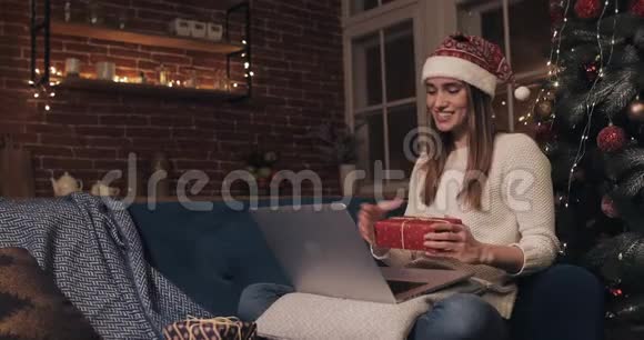 微笑的白种人年轻迷人的女孩戴着圣诞帽坐在圣诞树附近的沙发上做女人视频的预览图