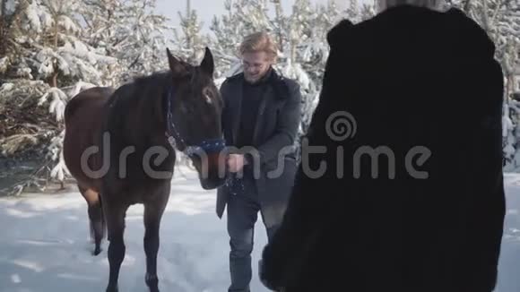 漂亮的女孩走到那家伙身边拥抱他英俊的年轻人挽着马缰站在后台视频的预览图