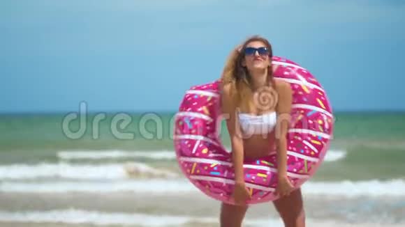 女人玩得开心笑着和粉红色的甜甜圈跳舞穿着比基尼的女孩在海滩上戴着太阳镜美丽性感的女人视频的预览图