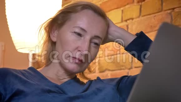 一位年长的白种人妇女在笔记本电脑上观看一场演出她聚精会神地坐在室内的沙发上拍摄特写镜头视频的预览图