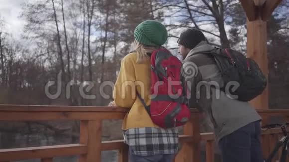 两个穿着夹克和帽子的快乐游客站在河边的桥上交谈享受大自然休假视频的预览图
