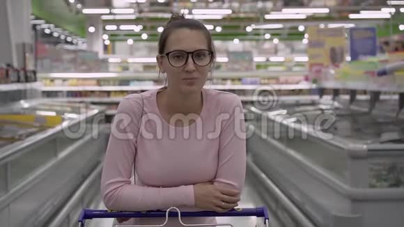 一个带眼镜的年轻孕妇站在超市里靠在手推车上做杂货店购物的女人视频的预览图