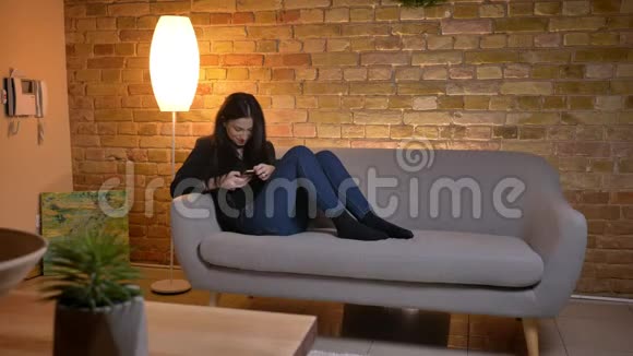 年轻的黑发白种人女孩坐在沙发上在家庭氛围中愉快地观看智能手机视频的预览图