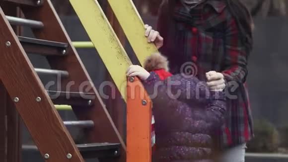妈妈帮助她幸福的女儿3岁至4岁的女孩在明亮多彩的操场上爬滑梯视频的预览图