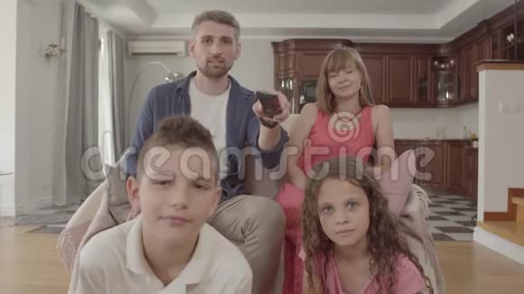 描绘了快乐的年轻家庭一起坐在家里看电视父亲拿着遥控器开关视频的预览图