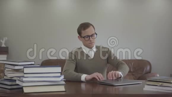 肖像可爱的书呆子戴着眼镜坐在办公室的木桌旁许多书都在桌子上可爱的男人视频的预览图